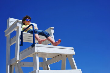 Cute little boy in sunglasses sitting on a lifeguard tower on the beach