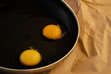 Two raw eggs with bright yellow yolks and transparent whites with a clots in a black frying pan on paper. The concept of  cooking a simple dish for breakfast. Close-up