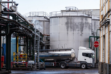 Tank truck loading in a factory next to some tanks and under pipes that give off steam.