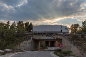 Fototapeta premium Truck with refrigerated semi-trailer passing over the top of a bridge over another road at sunset.