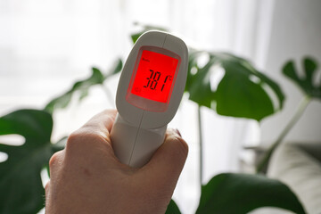 A female hand holding a forehead thermometer for body temperature showing 38,1 degrees, red