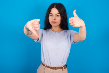 young beautiful tattooed girl wearing blue t-shirt standing against blue background showing thumbs up and thumbs down, difficult choose concept