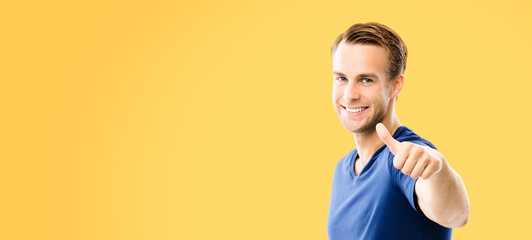 Portrait of happy smiling man in blue casual smart clothing, showing thumb up gesture, isolated over orange yellow color background. Male model at studio. Copy space area for some sign text.