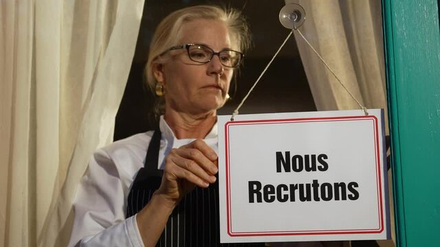 Female shop owner putting Help Wanted sign in French on glass door. Woman staff chef of restaurant advertising for hiring staff.