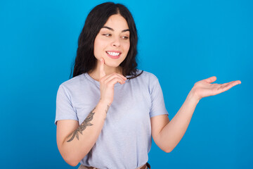 Funny young beautiful tattooed girl wearing blue t-shirt standing against blue background holding open palm new product. I wanna buy it!