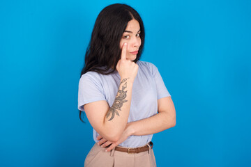 young beautiful tattooed girl wearing blue t-shirt standing against blue background Pointing to the eye watching you gesture, suspicious expression.