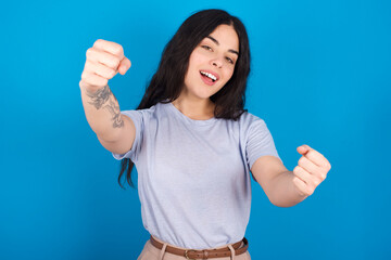 young beautiful tattooed girl wearing blue t-shirt standing against blue background  imagine steering wheel helm rudder passing driving exam good mood fast speed