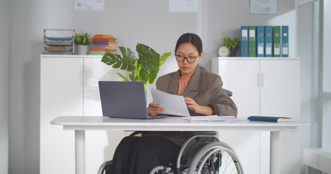 Young pretty disabled woman in wheelchair working on laptop computer at office.