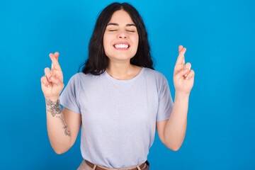 young beautiful tattooed girl wearing blue t-shirt standing against blue background gesturing finger crossed smiling with hope and eyes closed. Luck and superstitious concept.
