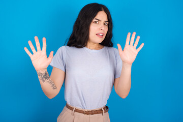 young beautiful tattooed girl wearing blue t-shirt standing against blue background afraid and terrified with fear expression stop gesture with hands, shouting in shock. Panic concept.