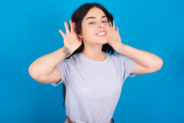 young beautiful tattooed girl wearing blue t-shirt standing against blue background Trying to hear both hands on ear gesture, curious for gossip. Hearing problem, deaf