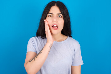 Fototapeta na wymiar Shocked young beautiful tattooed girl wearing blue t-shirt standing against blue wall looks with great surprisment being very stunned, astonished with unexpected news, Facial expressions concept.
