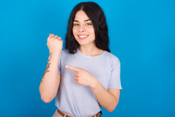 Fototapeta premium young beautiful tattooed girl wearing blue t-shirt standing against blue background In hurry pointing to wrist watch, impatience, looking at the camera with relaxed expression
