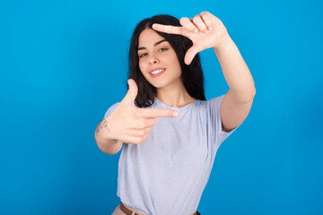 Fototapeta premium young beautiful tattooed girl wearing blue t-shirt standing against blue background making finger frame with hands. Creativity and photography concept.