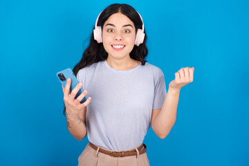 Positive young beautiful tattooed girl wearing blue t-shirt standing against blue background holds modern cell phone connected to headphones, clenches fist from good emotions, exclaims with joy,