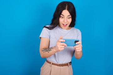 Portrait of an excited young beautiful tattooed girl wearing blue t-shirt standing against blue background playing games on mobile phone.