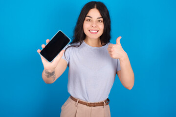 Portrait young beautiful tattooed girl wearing blue t-shirt standing against blue background holding in hands cell showing giving black screen thumb up