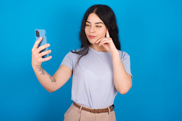 young beautiful tattooed girl wearing blue t-shirt standing against blue background smiling and...