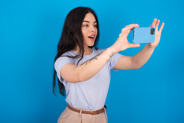Fototapeta premium young beautiful tattooed girl wearing blue t-shirt standing against blue background taking a selfie to post it on social media or having a video call with friends.
