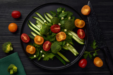 A black round plate of fresh vegetables stands on a black wooden table with a black knife, tomatoes and broccoli nearby.