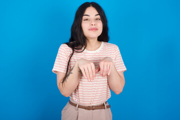 Fototapeta premium young beautiful tattooed girl wearing pink striped t-shirt standing against blue background makes bunny paws and looks with innocent expression plays with her little kid