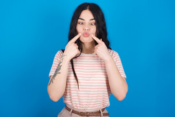 young beautiful tattooed girl wearing pink striped t-shirt standing against blue background crosses eyes and makes fish lips funny grimace