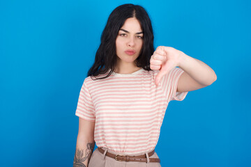 Fototapeta premium young beautiful tattooed girl wearing pink striped t-shirt standing against blue background looking unhappy and angry showing rejection and negative with thumbs down gesture. Bad expression.