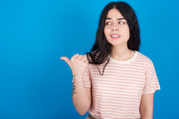 young beautiful tattooed girl wearing pink striped t-shirt standing against blue background points away and gives advice demonstrates advertisement