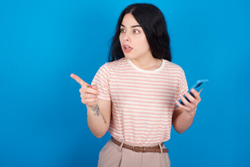 Stunned young beautiful tattooed girl wearing pink striped t-shirt standing against blue background points sideways right copy space, recommends product, sees astonishing thing