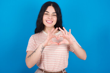 Serious young beautiful tattooed girl wearing pink striped t-shirt standing against blue background keeps hands crossed stands in thoughtful pose concentrated somewhere