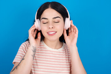 Fototapeta premium Pleased young beautiful tattooed girl wearing pink striped t-shirt standing against blue enjoys listening pleasant melody keeps hands on stereo headphones closes eyes. Spending free time with music