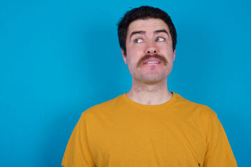 Fototapeta premium Coquettish young handsome Caucasian man with moustache wearing orange t-shirt against blue background smiling happily, blinking at camera in a playful manner, flirting with you.