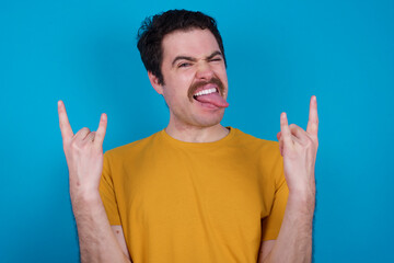 Naklejka premium young handsome Caucasian man with moustache wearing orange t-shirt against blue background making rock hand gesture and showing tongue