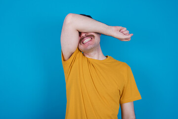 young handsome Caucasian man with moustache wearing orange t-shirt against blue background covering eyes with arm smiling cheerful and funny. Blind concept.