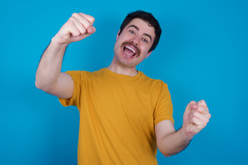 young handsome Caucasian man with moustache wearing orange t-shirt against blue background  imagine steering wheel helm rudder passing driving exam good mood fast speed