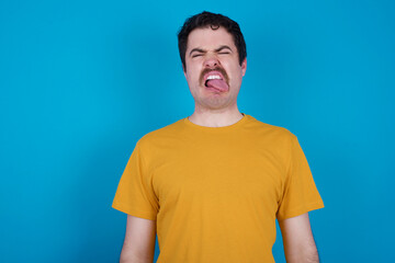 young handsome Caucasian man with moustache wearing yellow t-shirt against blue background sticking tongue out happy with funny expression. Emotion concept.