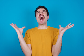 Fototapeta premium young handsome Caucasian man with moustache wearing yellow t-shirt against blue background crazy and mad shouting and yelling with aggressive expression and arms raised. Frustration concept.