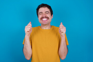 young handsome Caucasian man with moustache wearing yellow t-shirt against blue background being excited for success with raised arms and closed eyes celebrating victory. Winner concept.