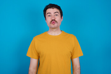 Shot of pleasant looking young handsome Caucasian man with moustache wearing yellow t-shirt against blue background, pouts lips, looks at camera, Human facial expressions