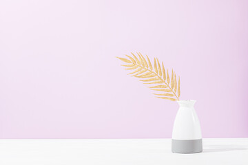 Golden palm leaf in vase on white table against lilac wall