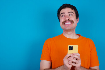 young handsome Caucasian man with moustache wearing orange t-shirt against blue background holding in hands showing new cell