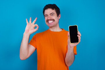young handsome Caucasian man with moustache wearing orange t-shirt against blue background holding...