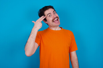 young handsome Caucasian man with moustache wearing orange t-shirt against blue background foolishes around shoots in temple with fingers makes suicide gesture. Funny model makes finger gun pistol