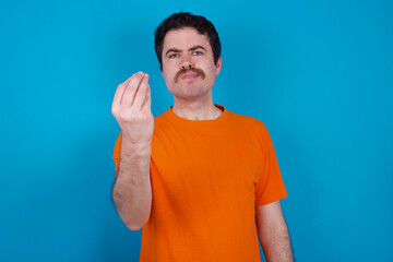 young handsome Caucasian man with moustache wearing orange t-shirt against blue background Doing Italian gesture with hand and fingers confident expression - obrazy, fototapety, plakaty