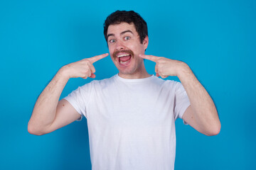 Cheerful young handsome Caucasian man with moustache wearing white t-shirt against blue background demonstrating hairdo