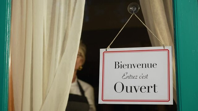 Female shop owner turning ouvert ferme sign on french cafe glass door. Woman staff chef of restaurant turning open closed sign in french.