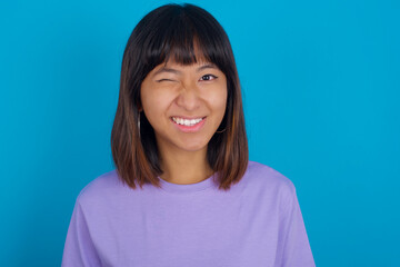 Coquettish Young beautiful asian girl wearing purple t-shirt against blue background smiling happily, blinking at camera in a playful manner, flirting with you.
