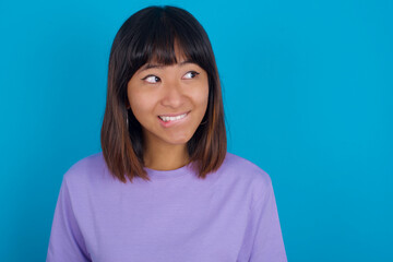 Fototapeta premium Amazed Young beautiful asian girl wearing purple t-shirt against blue background bitting lip and looking tricky to empty space.