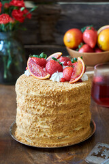 Layered honey cake garnished with strawberries, red orange and cream. Flowers, wooden background, side view.