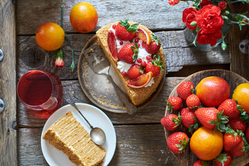 A piece of honey puff cake garnished with strawberries, red orange and cream. Flowers, wooden background, top view.